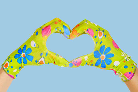 Hands in light green multi-colored gardening gloves folded in the shape of a heart isolated on a blue background. Hands in green gardening glovesの写真素材