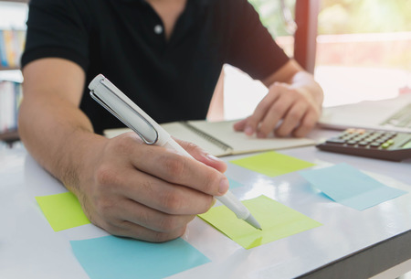 Businessman working and writing on sticky note paper on the table in office interior background.の写真素材