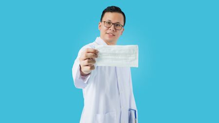 Portrait of Asian medical male Doctor holding a mask to prevent germs and standing isolated on blue...
