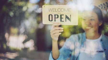Close up shot of woman hand turning open sign board on glass door in coffee shop and restaurant after coronavirus lockdown quarantine.Business crisis concept.の写真素材