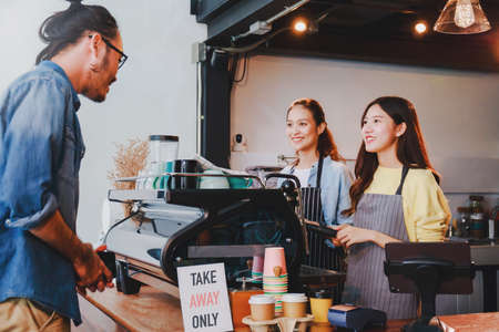 Young Asian woman barista wear apron making coffee cup served to customer at bar counter in coffee shop with smile face and takeaway orders only sign.Social distancing in situation of covid-19.の写真素材