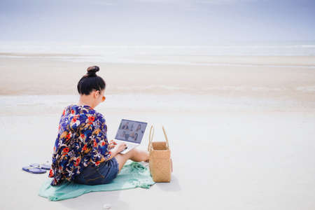 Asian happy woman sitting and using laptop meeting online on the beach by the sea background.Concept...