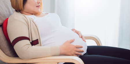 Pregnant Asian women wife sitting on sofa and use hand to touch the stomach at home background with copy space.Concept of maintaining health during a happy pregnancy.の写真素材