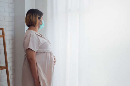 Pregnant Asian women wear a protective mask to prevent germs. Look out of the window During the quarantine at home for preventing coronavirus and covid-19 infection.の写真素材