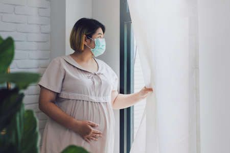 Pregnant Asian women wear a protective mask to prevent germs. Look out of the window During the quarantine at home for preventing coronavirus and covid-19 infection.の写真素材