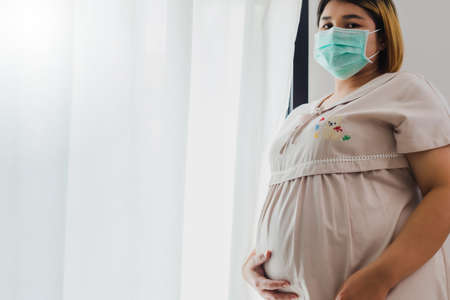 Pregnant Asian women wear a protective mask to prevent germs. Look out of the window During the quarantine at home for preventing coronavirus and covid-19 infection.の写真素材
