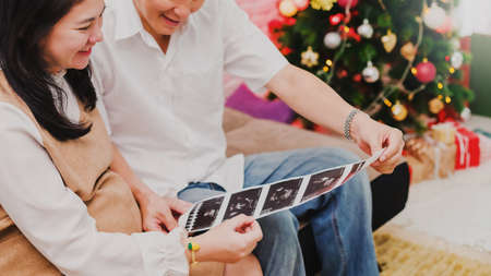 Pregnant Asian women wife sitting on sofa and see Ultrasound picture with husband at home background with Christmas tree.Concept of maintaining health during a happy pregnancy.の写真素材