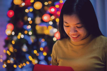 Asian woman open gift box with smiling face at home background with Christmas tree decoration for...