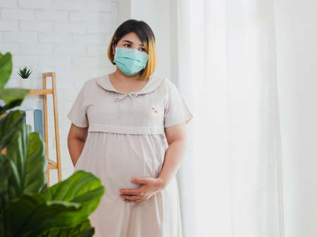 Pregnant Asian women wear a protective mask to prevent germs. Look out of the window During the quarantine at home for preventing coronavirus and covid-19 infection.の写真素材