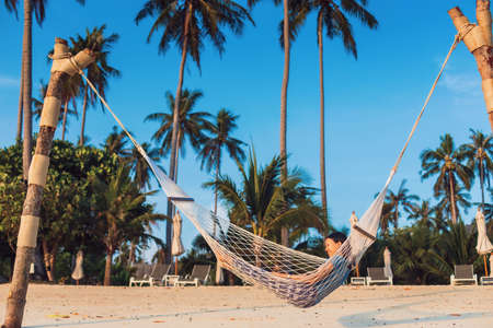 Asian traveler woman lying on the hammock on the beach in tropical resort hotel with Coconut tree and blue sky background.Travel in weekend holidays concept.の写真素材