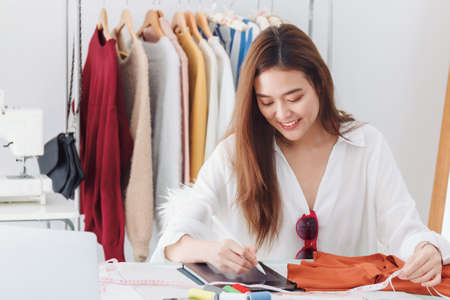Beautiful asian woman fashion designer sitting in the clothing store and studio. In front of the...