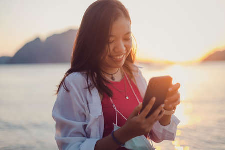 Asian traveler woman using smartphone online at the beach by the sea with sunset sky background.Concept of happy travel on vacation weekend.の写真素材
