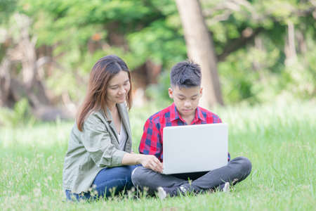 Cheerful mother and son sitting on the grass studying with laptop in the park, Outdoor learning conceptの写真素材