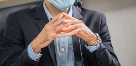 Close up of Man in suit sitting at a desk and clasp his hand, Young operator man with headset wear protection face maskの写真素材