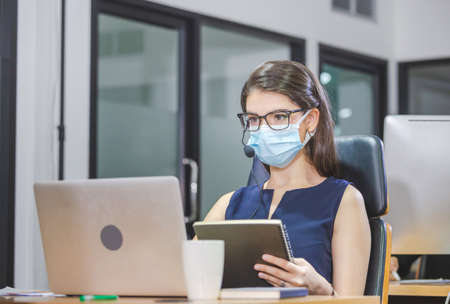 Young woman telephone operator with headset wear protection face mask against coronavirus, Customer service executive team working at officeの写真素材