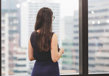 Businesswoman relaxing with holding cup of tea, woman standing near window looking out the window from a high floorの写真素材