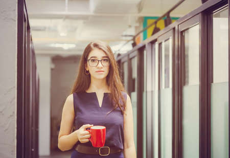 Beautiful woman with coffee walking in office, cheerful young business woman in office holding coffee cup and looking at camera.の写真素材