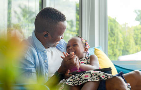 Cheerful african american father and daughter playing in living room, Cute little girl sitting on the sofa and playing on tabletの写真素材