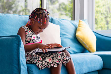 Cute little african american girl sitting on the sofa and playing on tablet in the living roomの写真素材