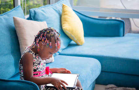 Little african american girl sitting on the sofa and playing on tablet in the living roomの写真素材