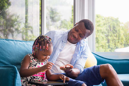 Cheerful african american father and daughter playing in living room, Cute little girl sitting on the sofa and playing on tabletの写真素材