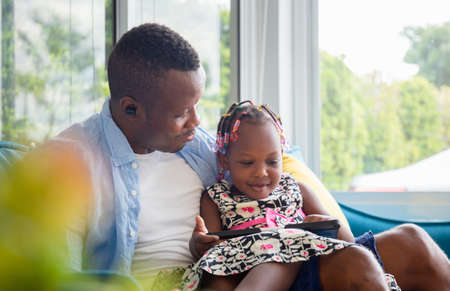 Cheerful african american father looking daughter playing in living room, Cute little girl sitting on the sofa and playing on tabletの写真素材