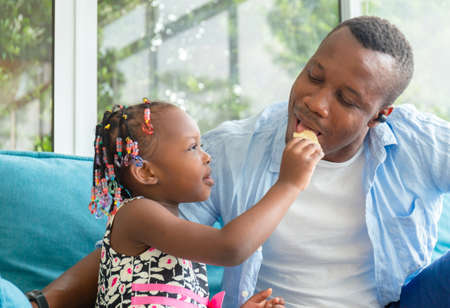 Cute little girl looking her dad eating snack, Cheerful african american father and daughter playing in living room, Happiness family conceptsの写真素材