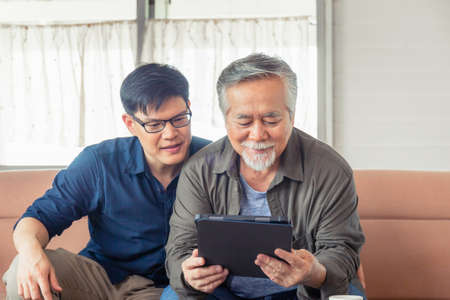 Happy senior asian father and adult son using tablet smartphone in living roomの写真素材