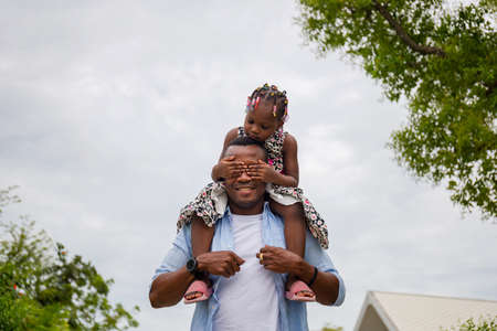 Happy father carrying daughter on shoulders, African american girl on the shoulders of his fatherの写真素材