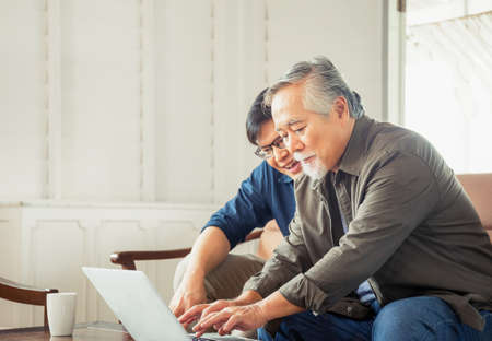 Happy senior asian father and adult son using Computer laptop talking on video call in living roomの写真素材