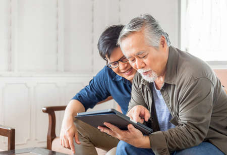 Happy senior asian father and adult son using tablet smartphone in living roomの写真素材