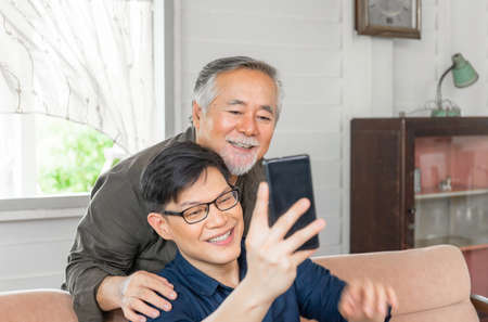 Happy senior asian father and adult son using smart phone talking on video call in living roomの写真素材