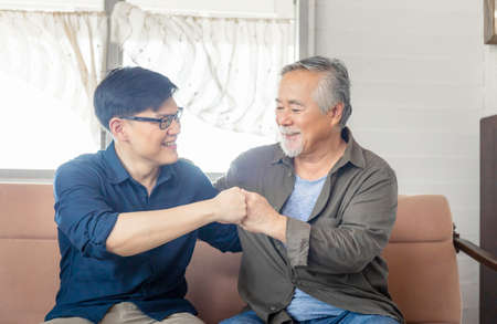 Cheerful senior asian father and middle aged son fists bumping, mature aged dad and millennial man sitting on couch in living room at home, Happiness Asian family conceptsの写真素材