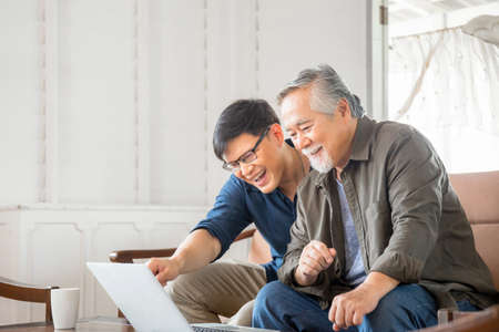 Happy senior asian father and adult son using laptop talking on video call in living roomの写真素材
