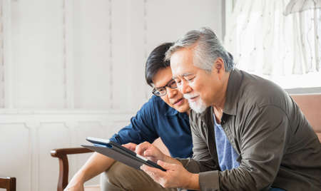 Happy senior asian father and adult son using tablet smartphone in living roomの写真素材