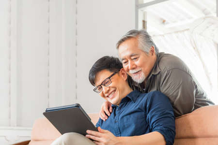 Happy senior asian father and adult son using tablet smartphone in living roomの写真素材