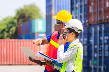 Worker man in hardhat and safety vest holding laptop and Female foreman talks on two-way radio control loading containers box from cargoの写真素材