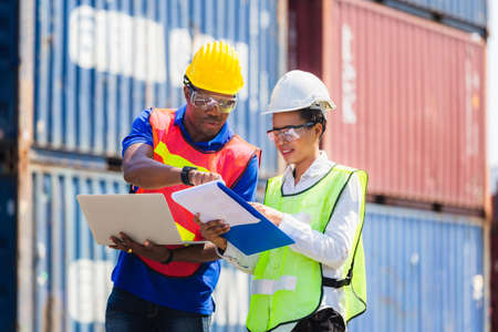 Female foreman safety vest using clipboard checklist and Worker man in hardhat holding laptop for control loading containers box from cargoの写真素材