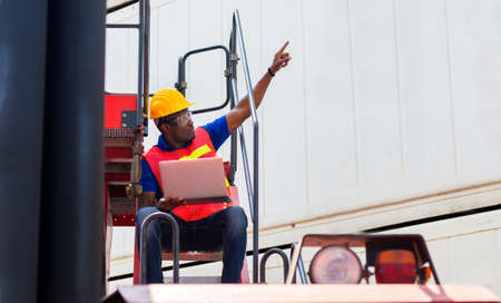 Worker man in hardhat and safety vest holding laptop on container stackers control loading containers box from cargoの写真素材