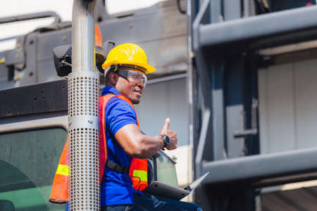 Worker in hardhat and safety vest holding laptop standing on container stackers control loading containers box from cargo, Smiling man and giving thumbs upの写真素材