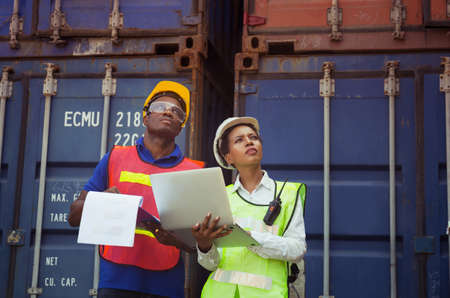 Worker man in hardhat and safety vest holding clipboard checklist and Female foreman using laptop control loading containers box from cargoの写真素材