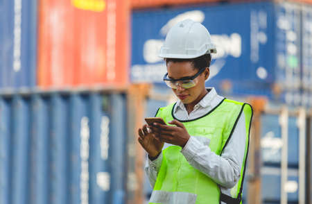 Female engineer in the hard hat uses mobile phone, Industrial worker using mobile smartphone in industry containers cargoの写真素材
