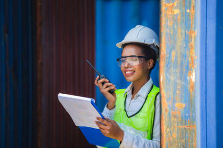 Woman Foreman in hardhat and safety vest holding clipboard checklist and talks on two-way radio control loading containers box from cargoの写真素材