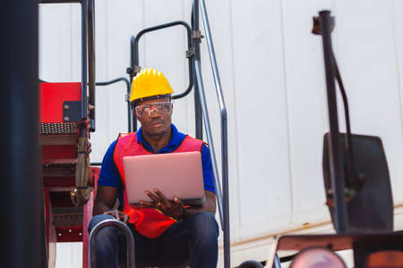 Worker man in hardhat and safety vest holding laptop standing on container stackers control loading...
