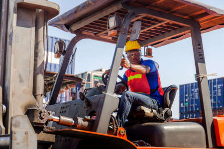 Worker man in hardhat and safety vest sitting in container stackers control loading containers box from cargoの写真素材