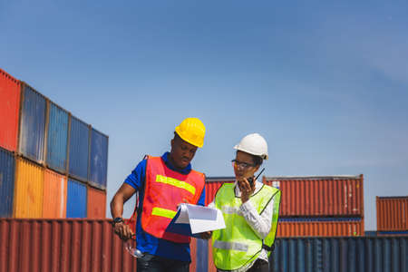 Worker man in hardhat and safety vest holding clipboard checklist and Female foreman talks on two-way radio control loading containers box from cargoの写真素材