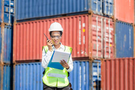 Woman Foreman in hardhat and safety vest holding clipboard checklist and talks on two-way radio control loading containers box from cargoの写真素材