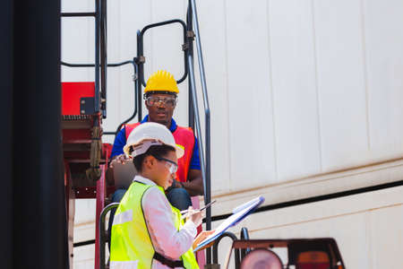 Female foreman safety vest using clipboard checklist and Worker man in hardhat holding laptop for control loading containers box from cargoの写真素材
