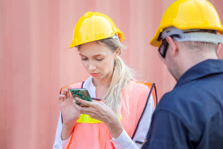 Engineer in the hard hat uses mobile phone, Industrial worker using mobile smartphone in industry containers cargoの写真素材