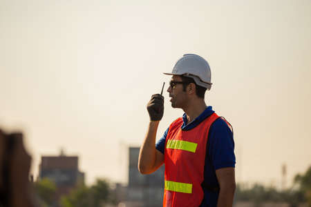 Foreman worker in hardhat and safety vest talks on two-way radio control loading containers box from cargoの写真素材